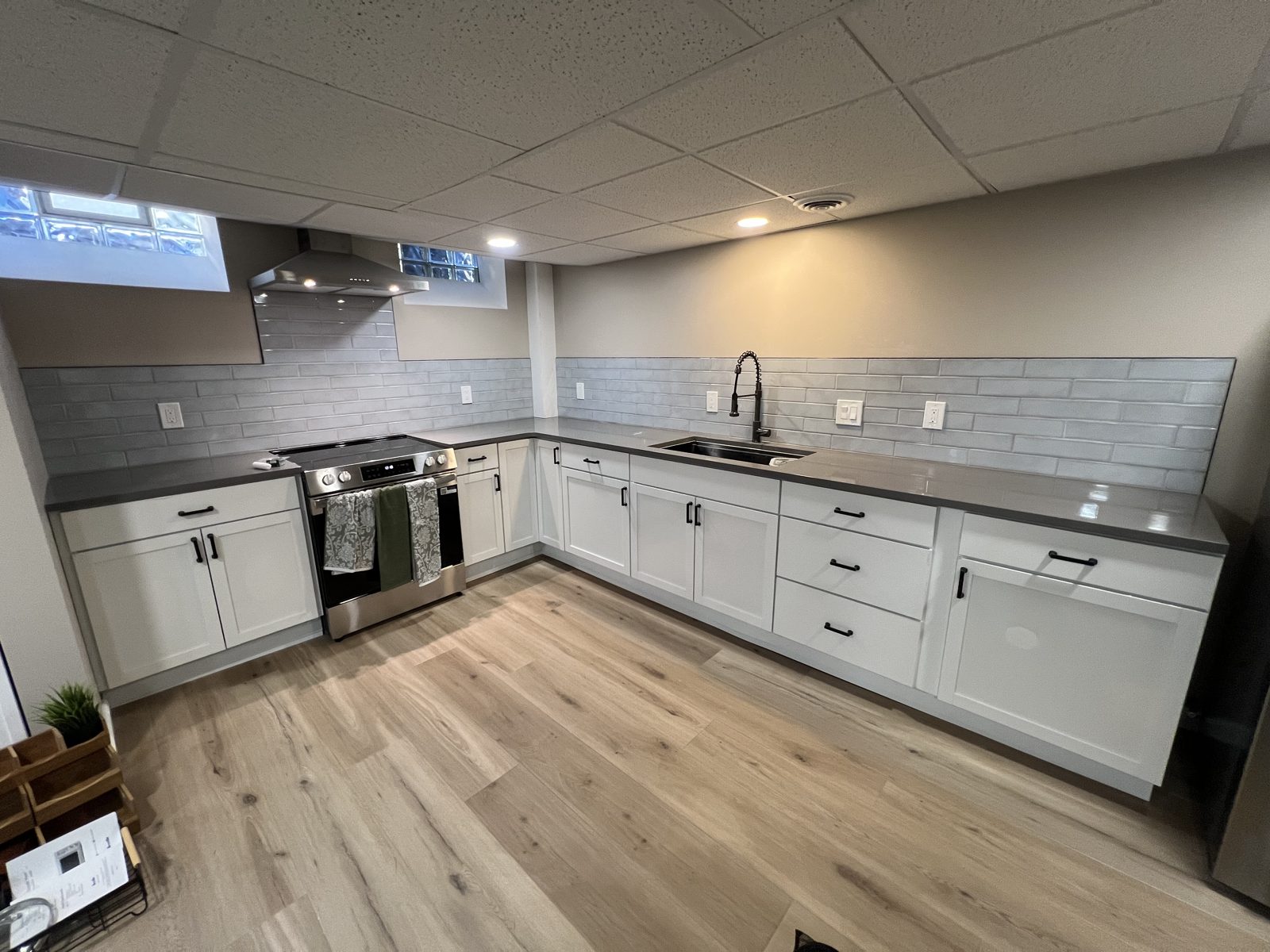 Glazed gray subway tile backsplash running the length of a basement kitchen and up behind the range hood, above white cabinets and gray quartz counters