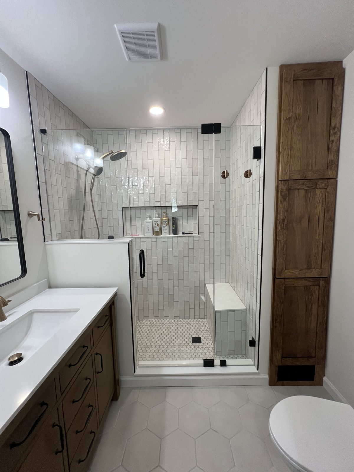 Bathroom renovated down to the studs, with a walk-in tiled shower, recessed niche, built-in bench and frameless glass, beside a wood vanity and a tall custom wood cabinet